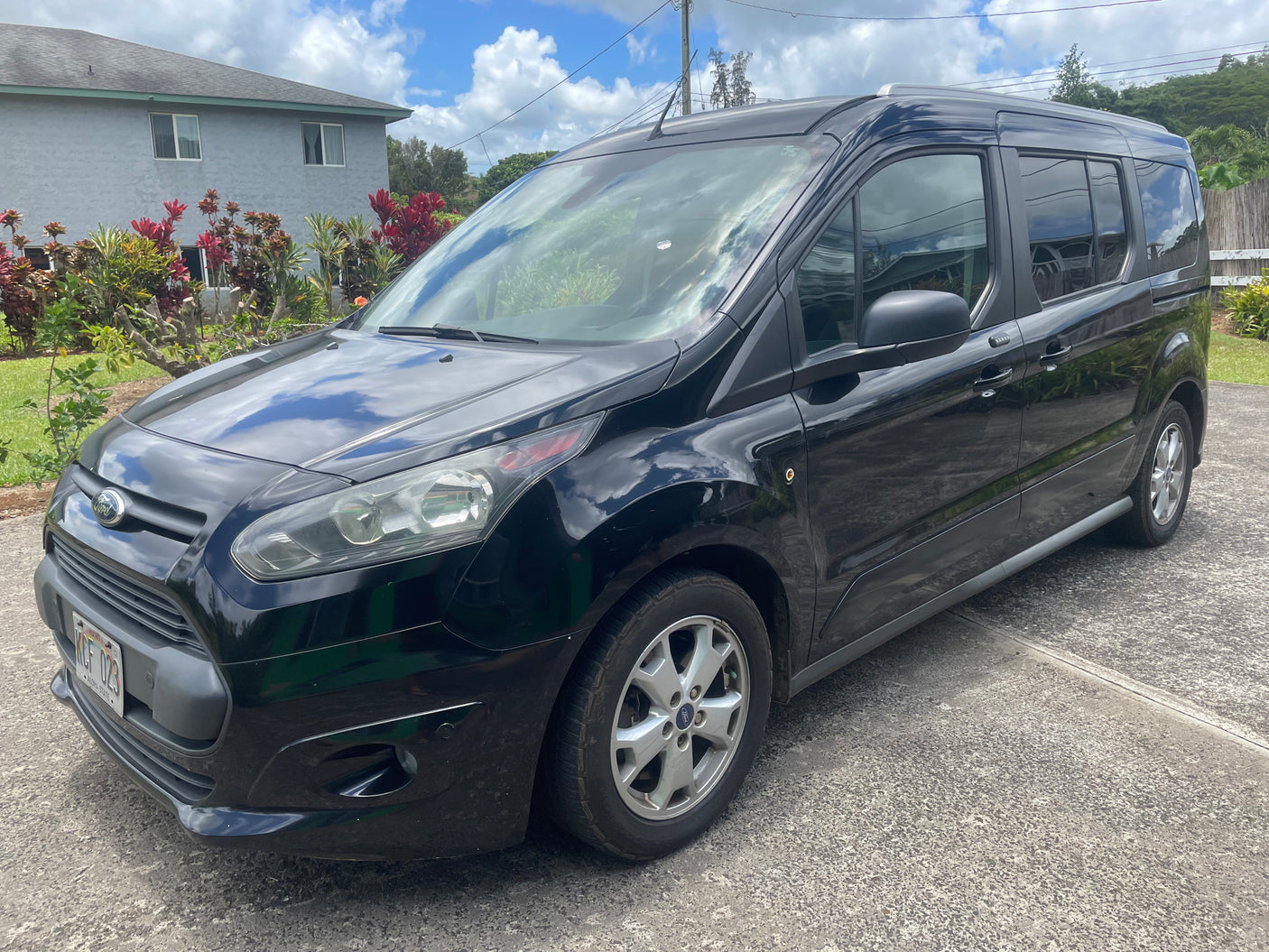 Black Ford Transit Connect passenger van on a driveway, ideal for a compact RV rental. Sunny day, house in background.