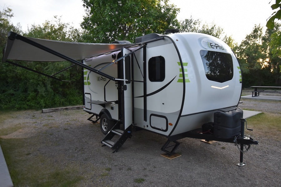 This white and lime green E-Pro RV rental is parked with its awning extended at a gravel campsite, perfect for outdoor trips.
