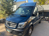 Dark blue Mercedes-Benz Sprinter camper van for RV rental, parked on a driveway in front of a house.