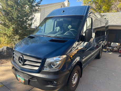 Dark blue Mercedes-Benz Sprinter camper van for RV rental, parked on a driveway in front of a house.
