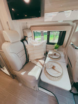 Inside a modern RV rental: a bright living area with dining table for two, comfortable seats, and a TV.