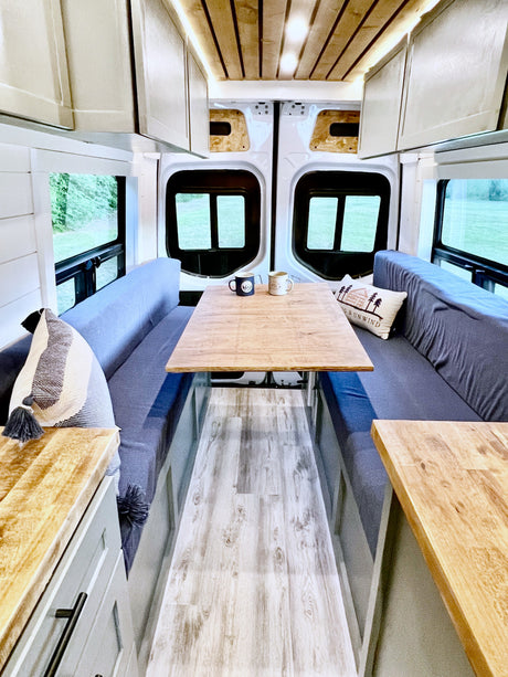 Modern rv rental interior with bright wood ceiling, white cabinetry, long gray benches, and a central wood dining table.