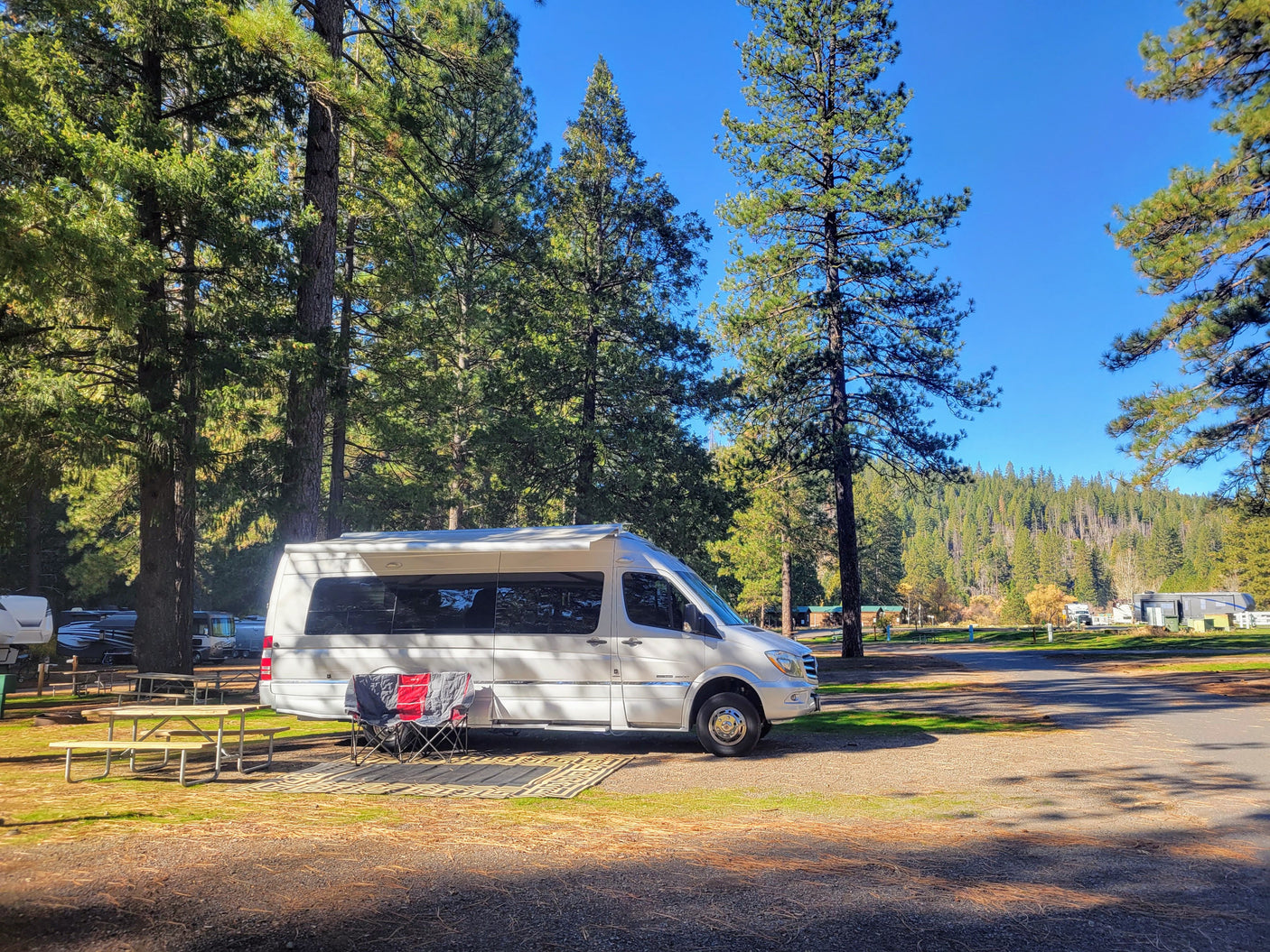 White RV rental parked at a sunny campground site, with a picnic table, chairs, and tall trees providing shade.