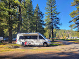 White RV rental parked at a sunny campground site, with a picnic table, chairs, and tall trees providing shade.