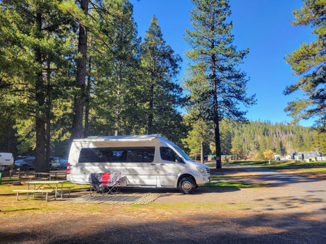 White RV rental parked at a sunny campground site, with a picnic table, chairs, and tall trees providing shade.