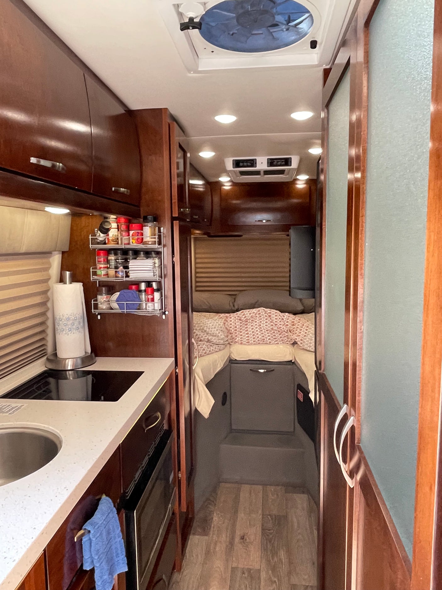 This RV rental interior showcases a compact kitchen with a sink, cooktop, and spice rack, leading to a cozy bedroom area.