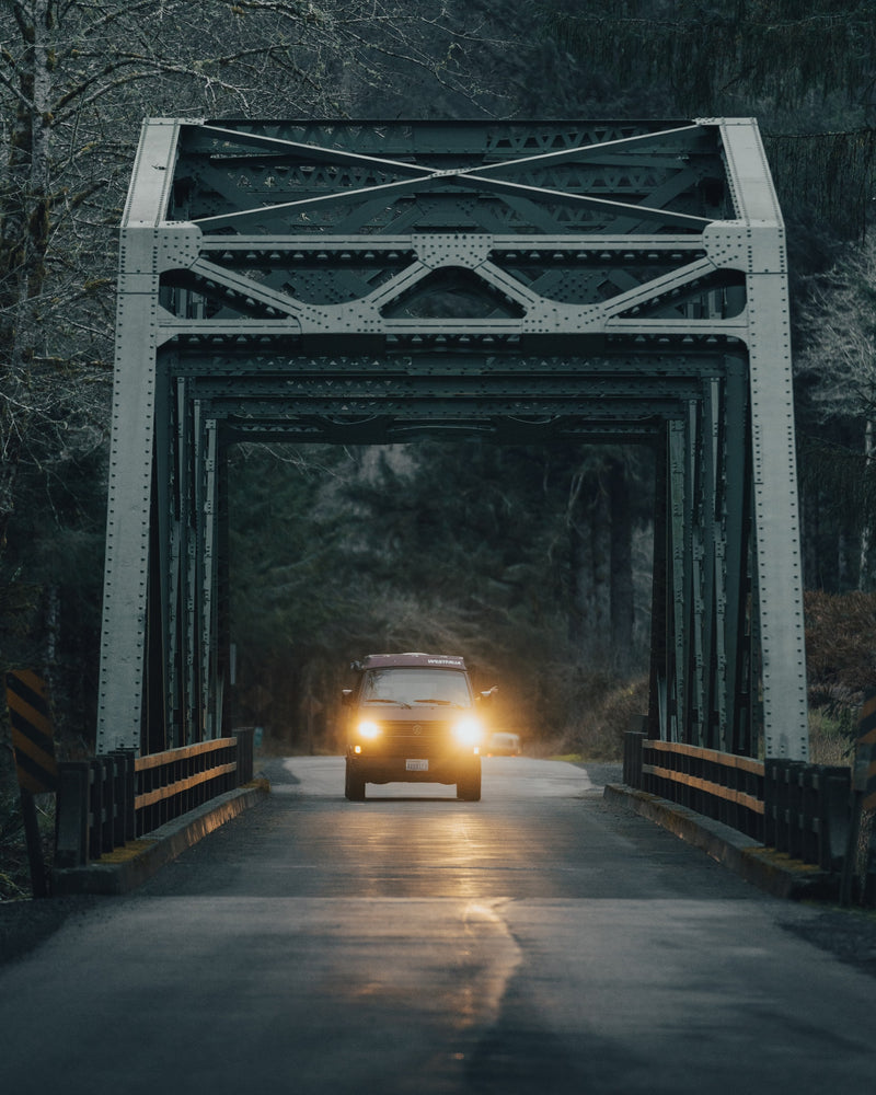 An RV rental adventure! A classic VW Westfalia camper van, headlights beaming, drives through a moody, metal truss bridge. — photo 7