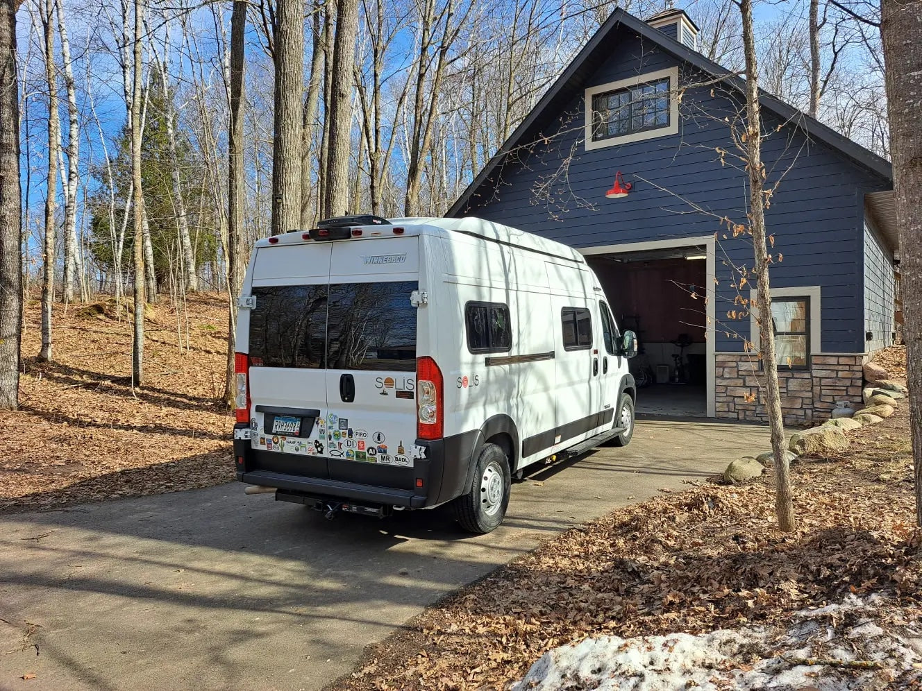 White Winnebago Solis RV rental parked in a driveway next to a blue house in a wooded area with bare trees and fallen leaves.