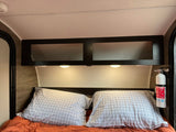 RV rental interior showing a bed with pillows, overhead cabinets, reading lights, and a visible fire extinguisher for safety.