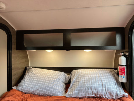 RV rental interior showing a bed with pillows, overhead cabinets, reading lights, and a visible fire extinguisher for safety.