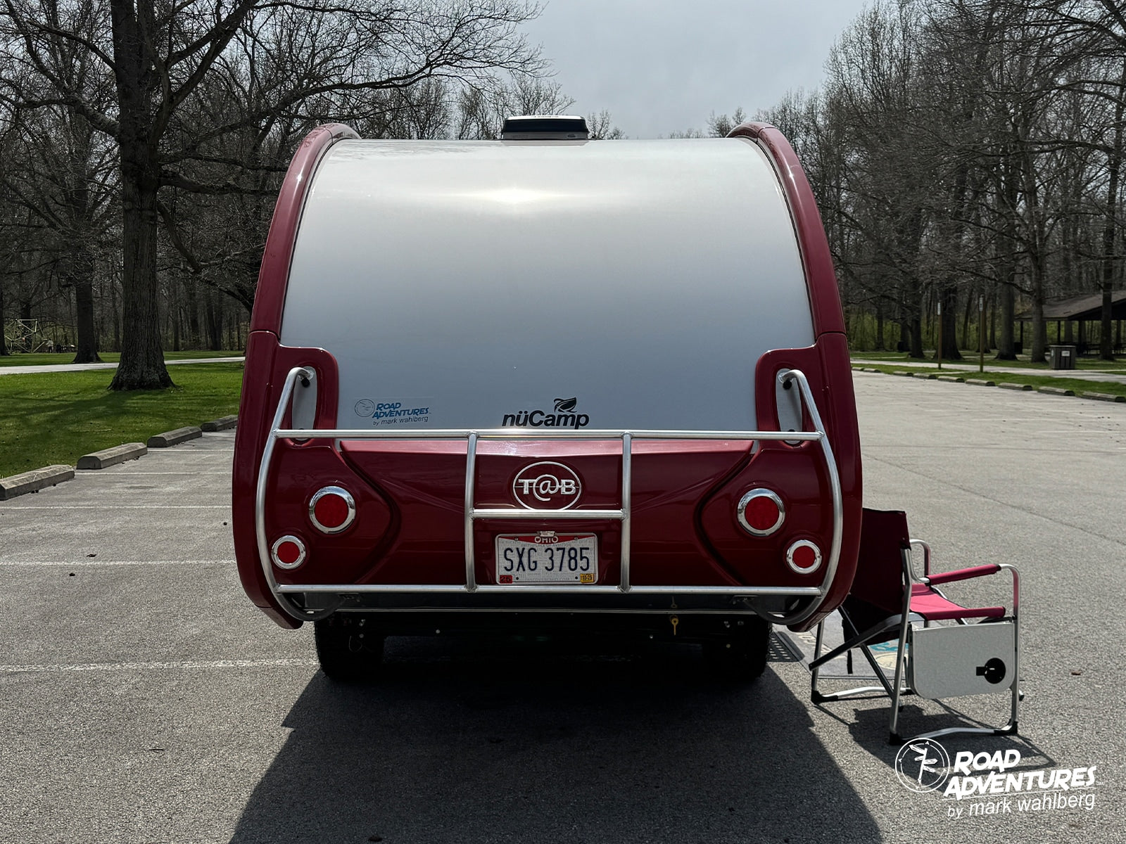Rear view of a red and white nüCamp T@B travel trailer, an ideal rv rental for park adventures. A camping chair sits nearby.