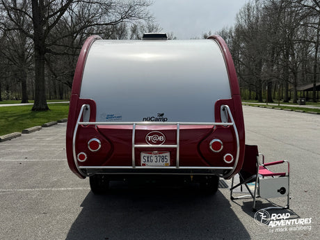 Rear view of a red and white nüCamp T@B travel trailer, an ideal rv rental for park adventures. A camping chair sits nearby.