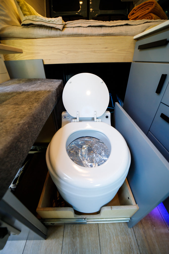 Compact RV rental interior with a pull-out portable toilet, lid open, revealing a foil-lined bowl. Ideal for van life adventures.