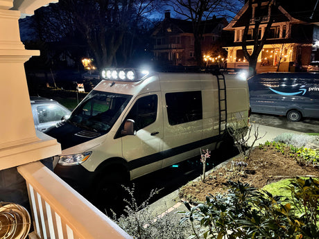 An adventure van, suitable for rv rental, with bright roof lights, parked at night with an Amazon delivery van nearby.