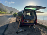 Red camper van with open rear hatch showing bed, parked on sandy shoulder by beach and mountains. Ideal rv rental for ocean camping.