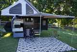This RV rental camper trailer is set up for outdoor adventure, featuring an awning, pop-top tent, chairs, and an outdoor kitchen.