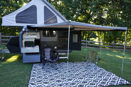 This RV rental camper trailer is set up for outdoor adventure, featuring an awning, pop-top tent, chairs, and an outdoor kitchen.