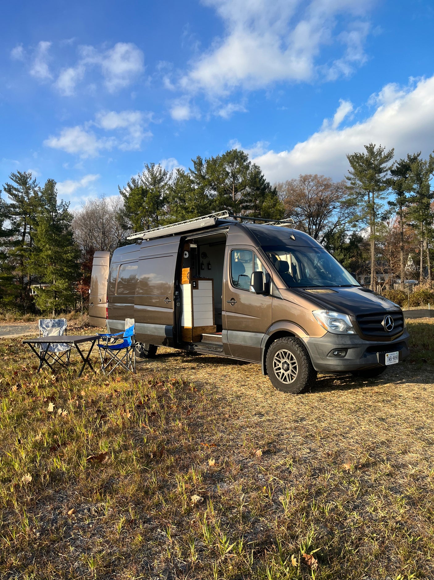Brown Mercedes-Benz Sprinter rv rental on sunny grass. Open side door reveals interior. Chairs & table set up.