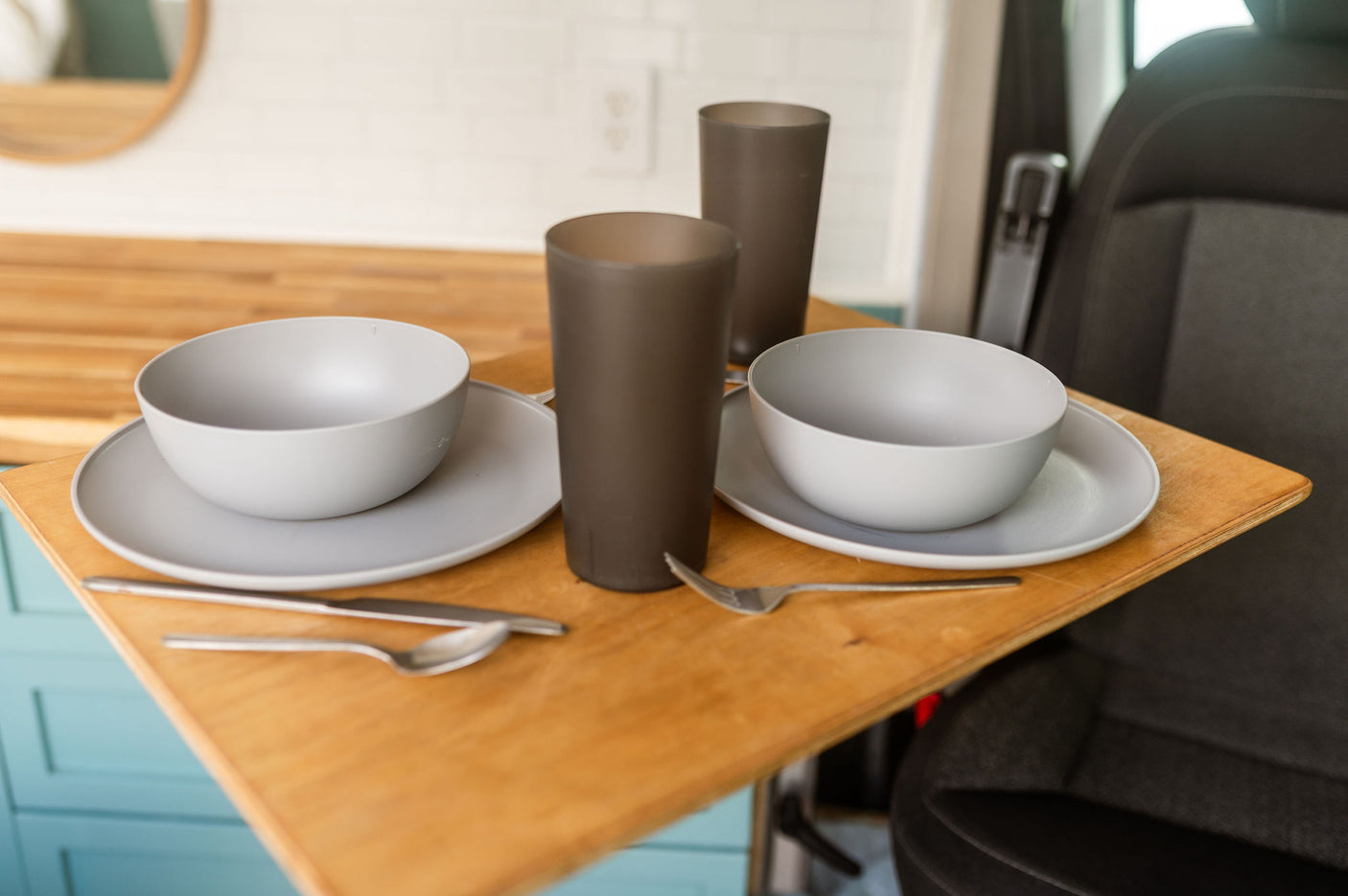 RV rental compact dining: light gray bowls, plates, dark tumblers, and silverware on a wooden table next to a vehicle seat.