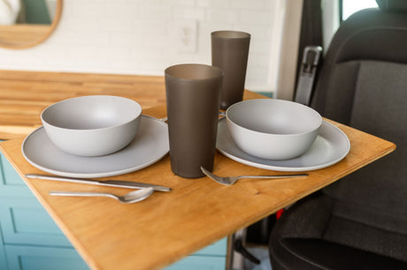 RV rental compact dining: light gray bowls, plates, dark tumblers, and silverware on a wooden table next to a vehicle seat.
