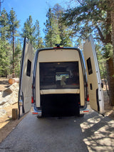 Rear view of an RV rental camper van with open doors, showing its cozy interior, parked in a sunny forest. Perfect for nature adventures.
