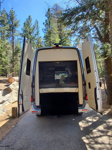 Rear view of an RV rental camper van with open doors, showing its cozy interior, parked in a sunny forest. Perfect for nature adventures.