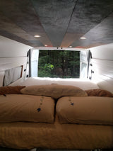 Relax in this cozy RV rental's bed, looking out at a tranquil forest.