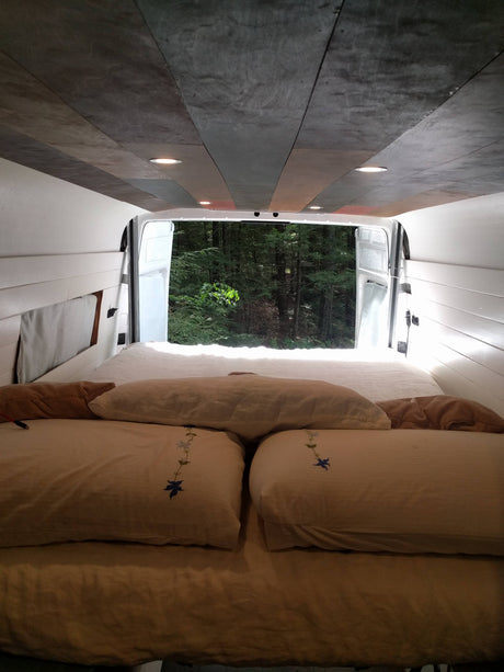 Relax in this cozy RV rental's bed, looking out at a tranquil forest.