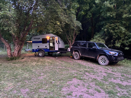RV rental: A compact Sunray Sport travel trailer & dark SUV parked at a wooded campsite, ideal for outdoor adventure.