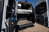 Inside a custom rv rental: rear view of a camper van's organized storage area with a bed, wood floor, and door panels.