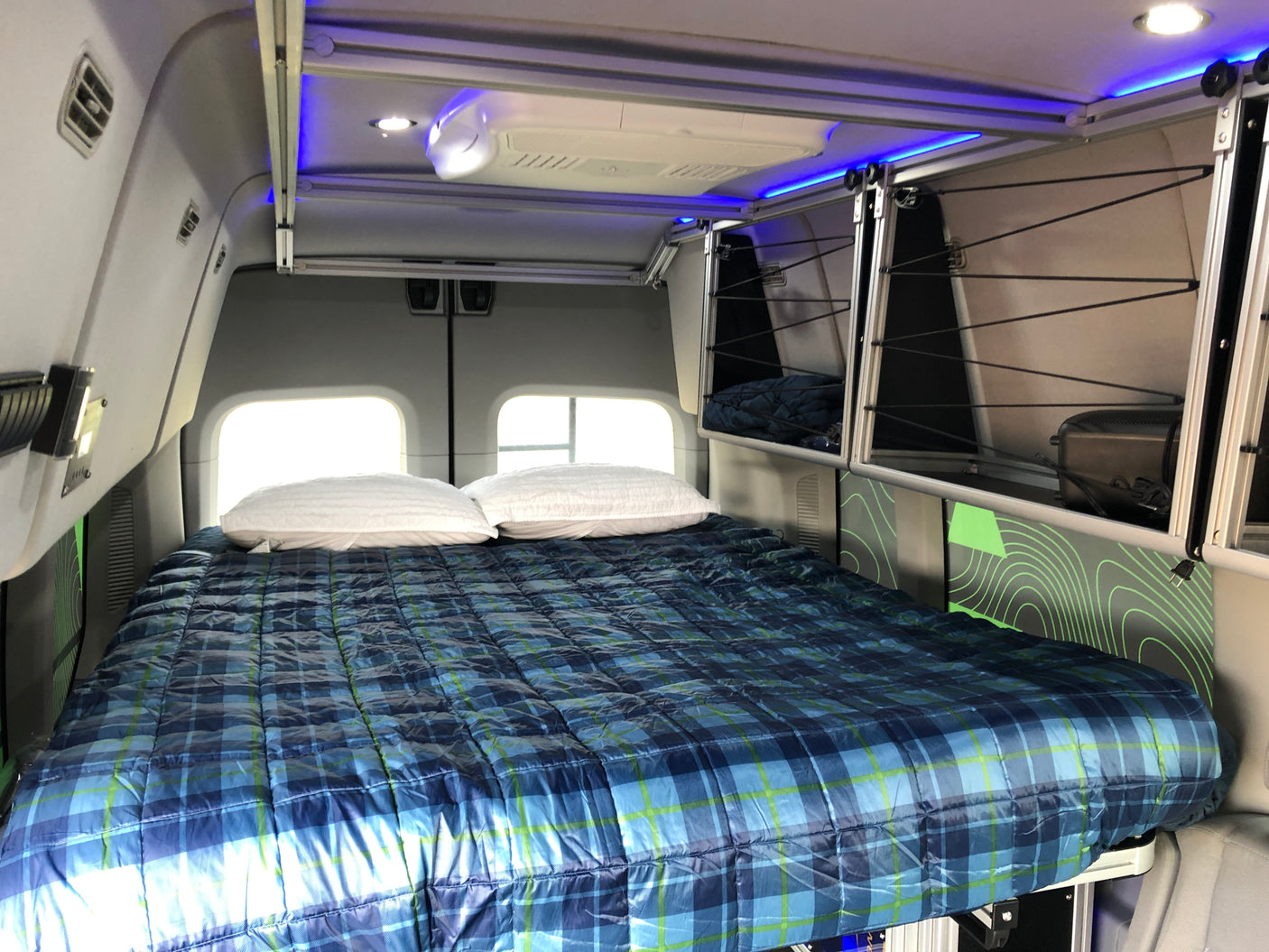Inside a modern **RV rental**: a plush bed with blue plaid comforter, ample overhead storage, and cool blue ambient lights.