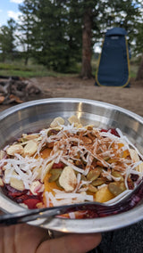 A nourishing outdoor meal in a metal bowl (nuts, fruit, coconut) at an RV rental site with a blue privacy tent.