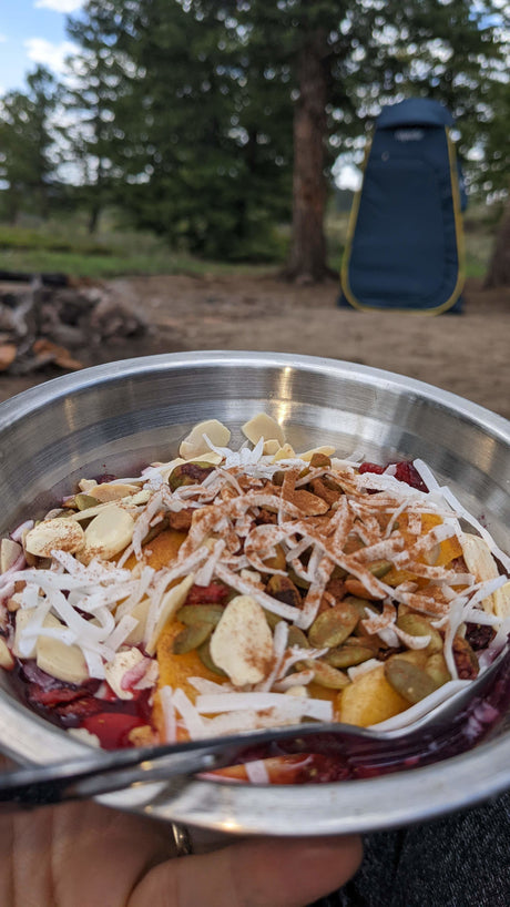 A nourishing outdoor meal in a metal bowl (nuts, fruit, coconut) at an RV rental site with a blue privacy tent.