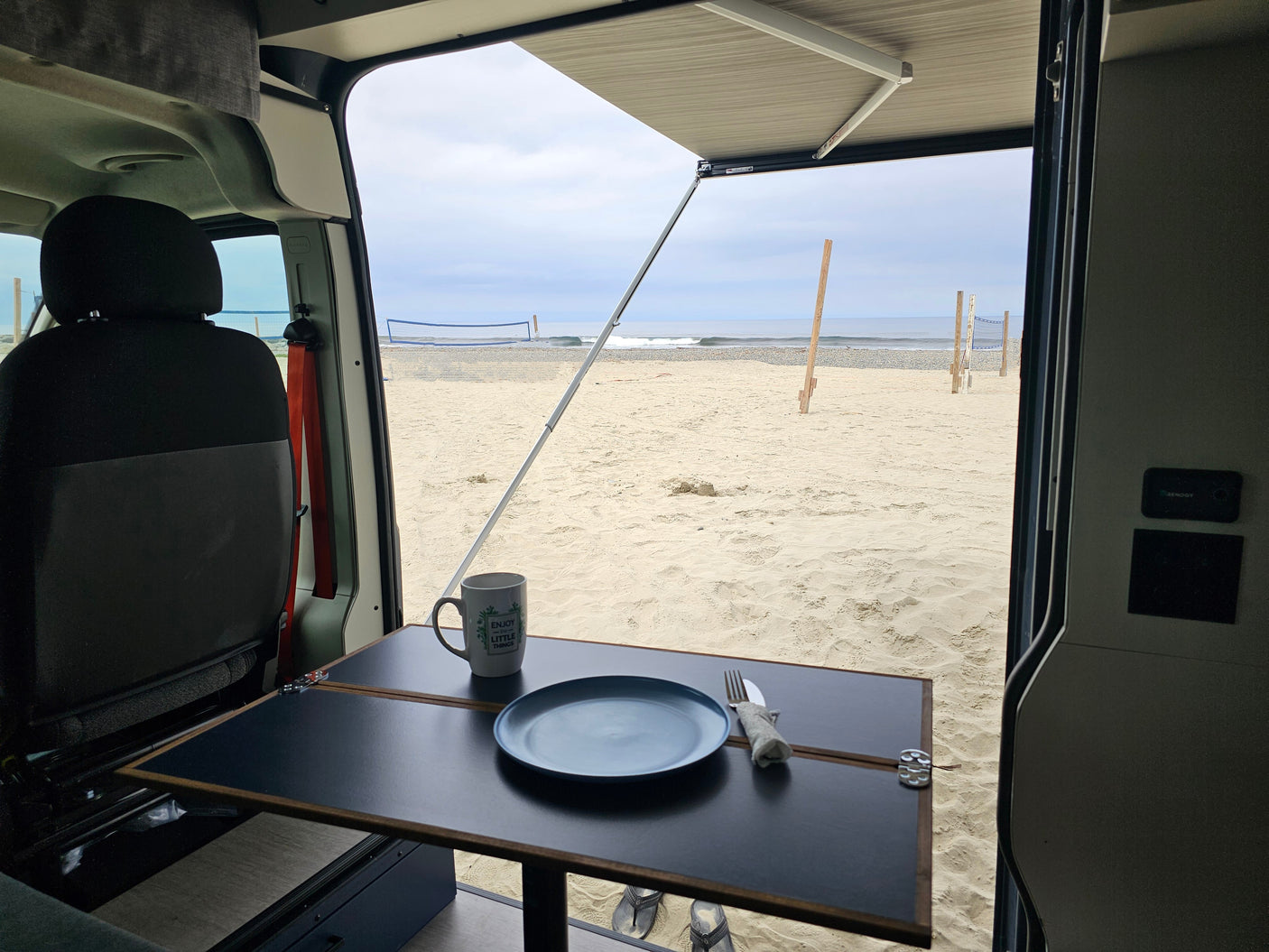Experience beach life in an RV rental! Inside a campervan, table set for breakfast with ocean & volleyball net view.