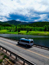 Grey RV rental on a road beside a scenic lake, framed by green forests and mountains under a dramatic sky.