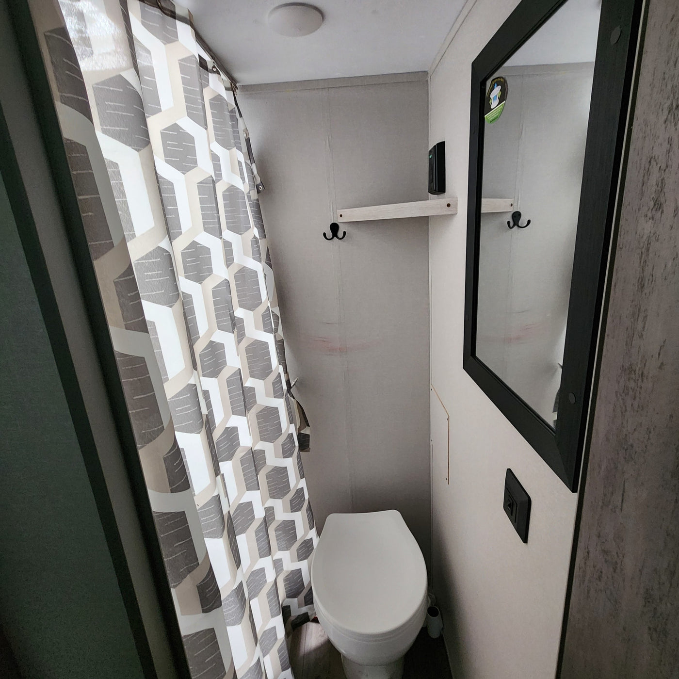 Compact RV rental bathroom features a white toilet, geometric gray/white shower curtain, mirror, and small shelf with hooks. — photo 8