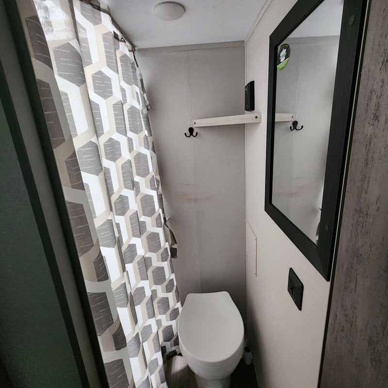 Compact RV rental bathroom features a white toilet, geometric gray/white shower curtain, mirror, and small shelf with hooks. — photo 8