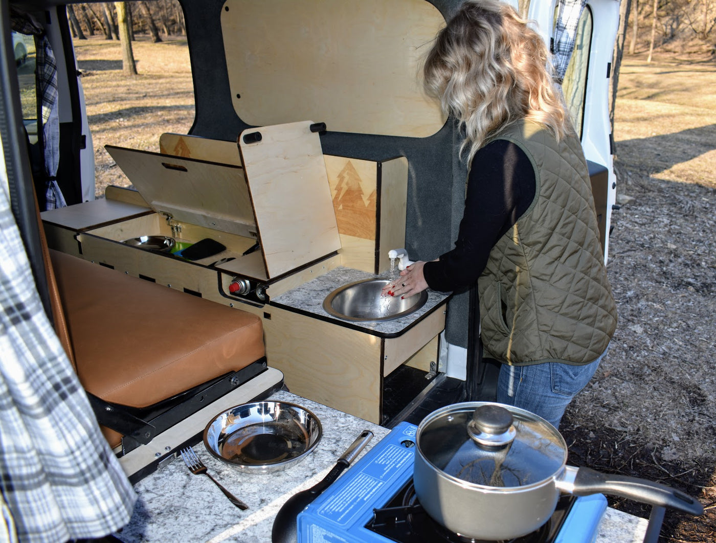 Inside an RV rental camper van: Woman uses the integrated sink, next to a portable stove and modular bed. — photo 3