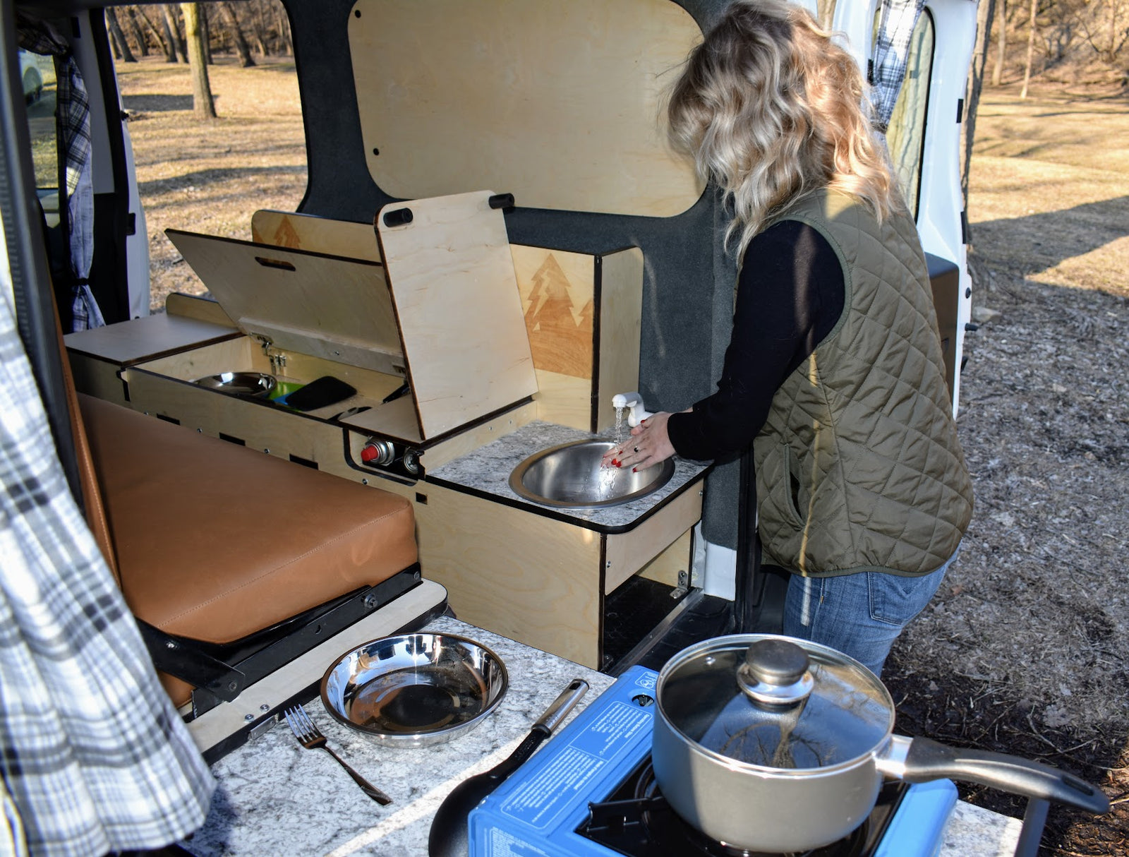 Inside an RV rental camper van: Woman uses the integrated sink, next to a portable stove and modular bed.