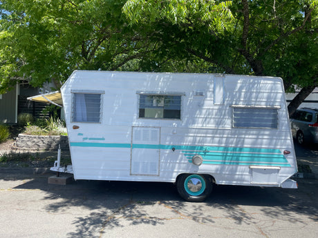 A charming white and teal vintage travel trailer, ideal for an rv rental, rests on a sunny street, shaded by green trees.