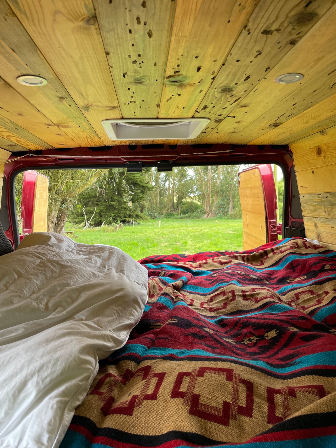 Inside a cozy RV rental: a bed with patterned blanket, wooden ceiling, and open rear doors revealing a serene green field.