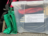 RV rental gear: Clear bin labeled with power cord, water hose, jumper cables, tire pump, and emergency supplies. Green tarp nearby.