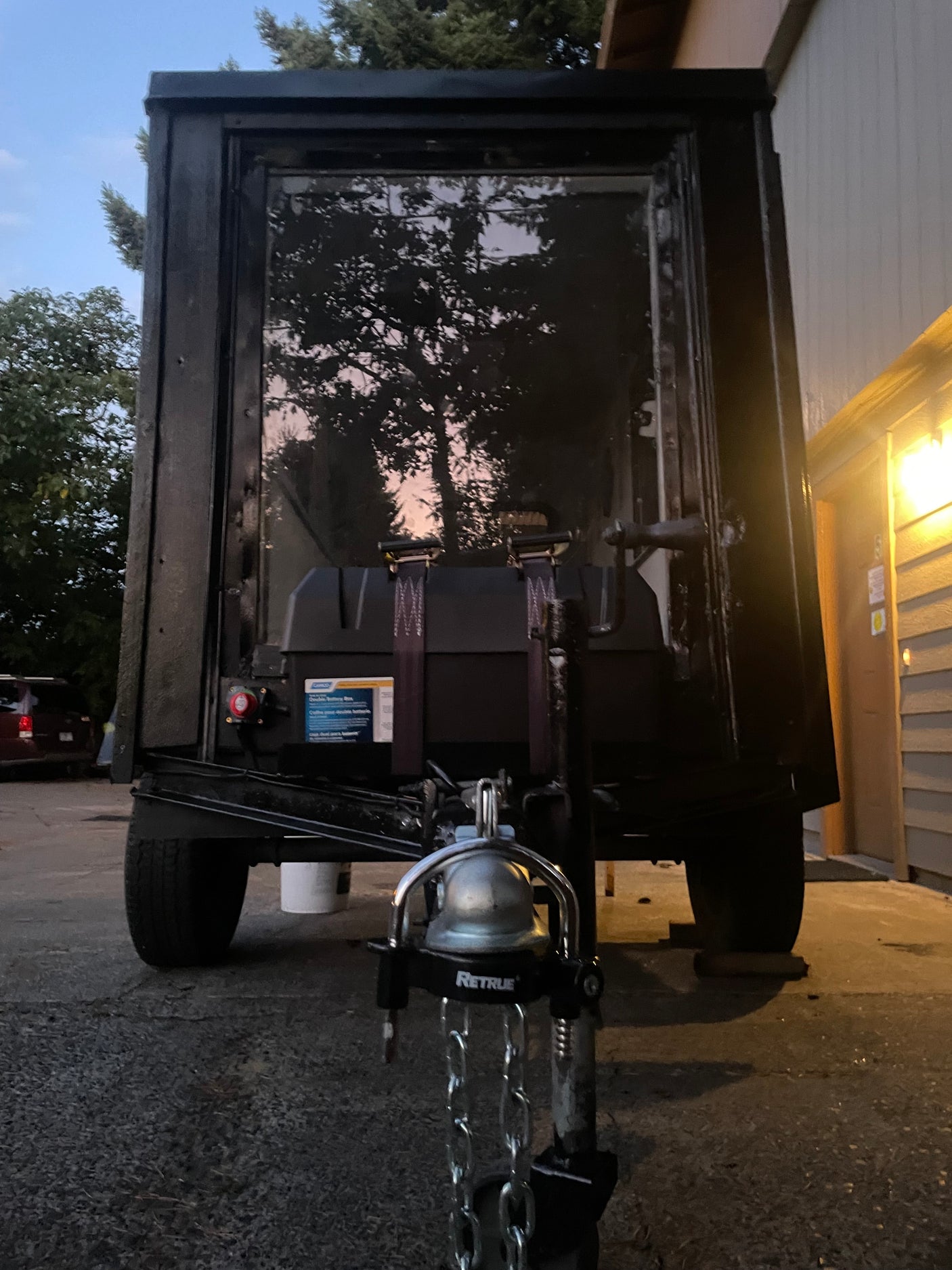 Back of a dark RV rental trailer with hitch, safety chains, & battery box. Window reflects trees & sky. Parked outdoors.