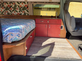 Inside this vibrant RV rental: a cozy bed with storage, compact kitchen with sink, and bright red cabinets, ready for adventure.