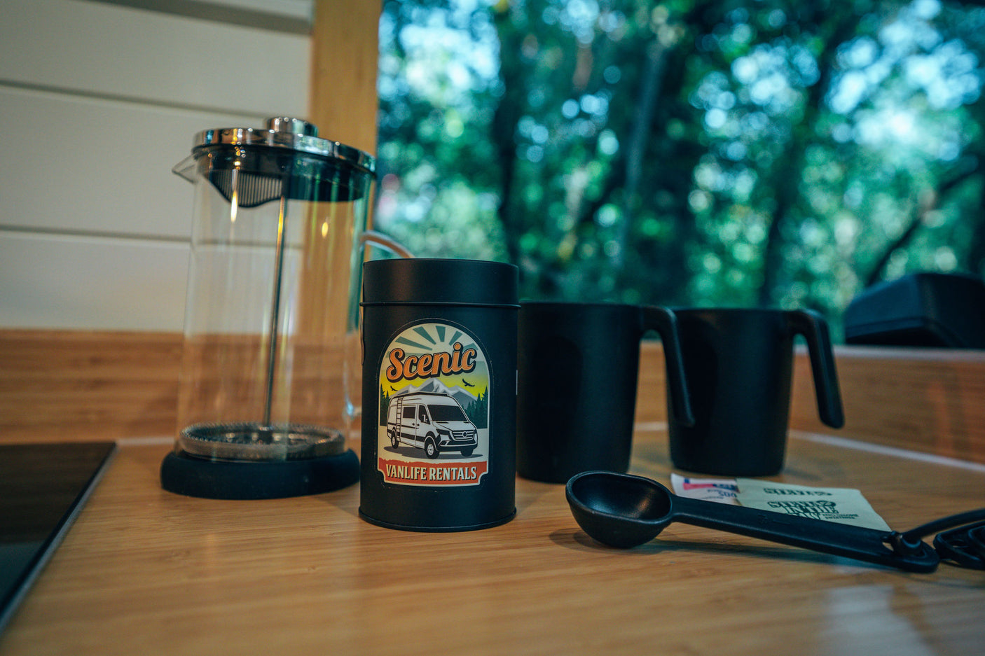 Coffee setup in an RV rental: French press, Scenic Vanlife Rentals canister, mugs, and spoon on a counter with a forest view.