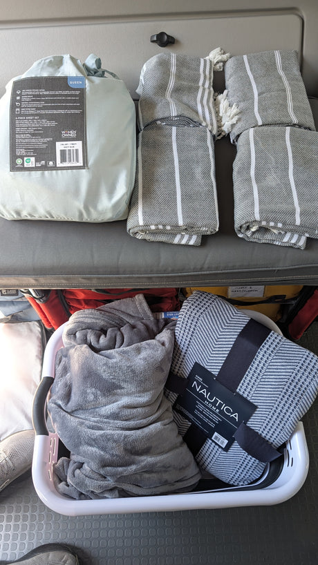 Preparing an RV rental with cozy bedding: a queen sheet set, striped blankets, and a plush blanket for travel comfort.