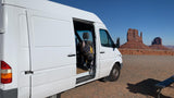 A white rv rental van, side door open, parked in a desert lot. Iconic red rock formations of Monument Valley rise under a blue sky.