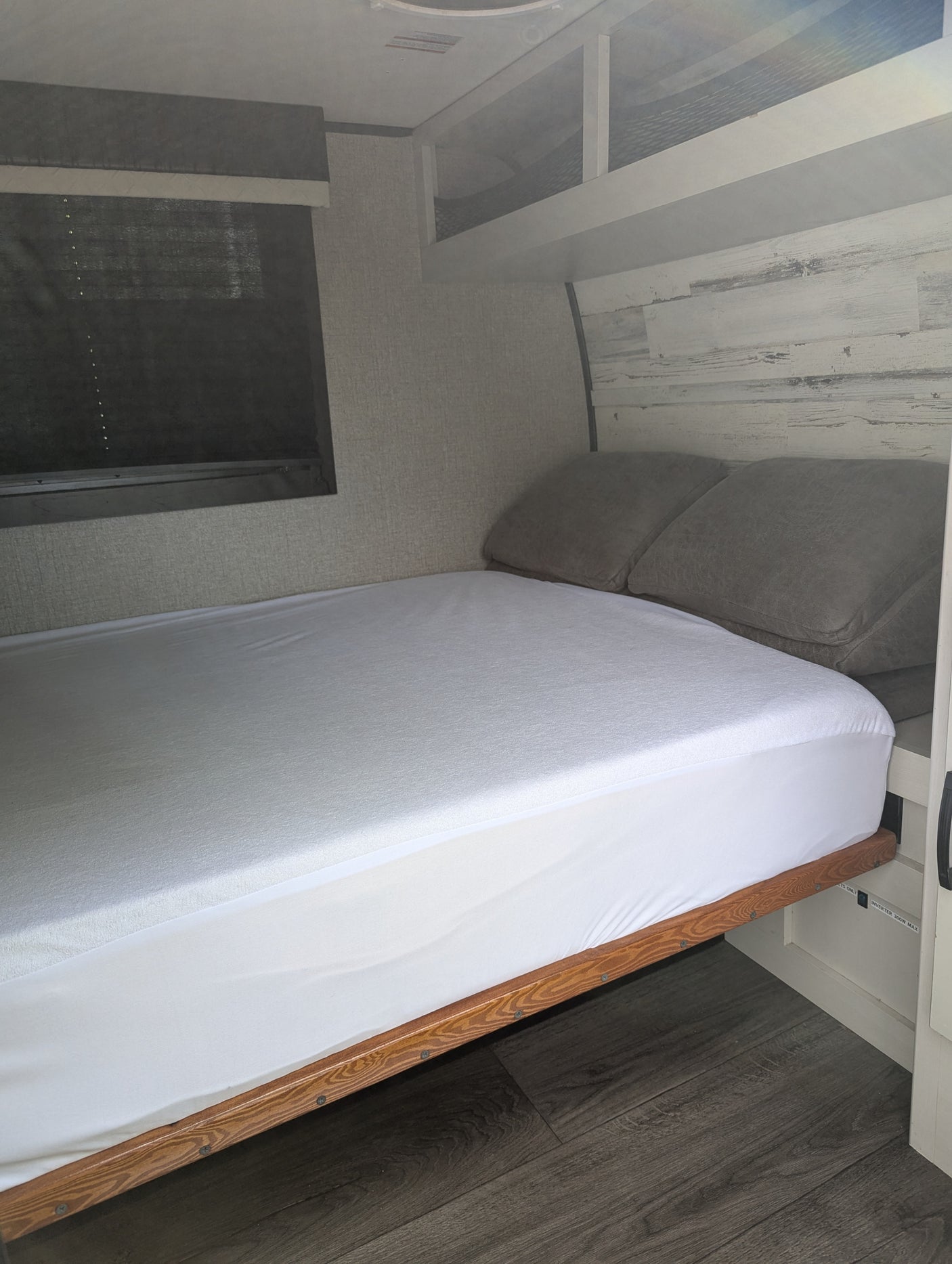 Modern RV rental bedroom features a fresh white bed, grey pillows, and stylish wood-look decor. Window with blinds.