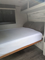 Modern RV rental bedroom features a fresh white bed, grey pillows, and stylish wood-look decor. Window with blinds.