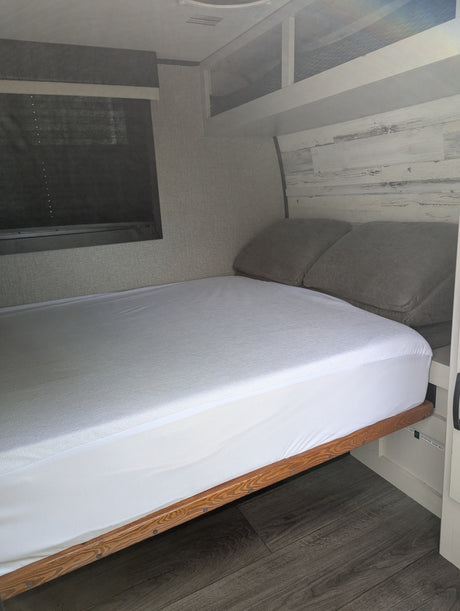 Modern RV rental bedroom features a fresh white bed, grey pillows, and stylish wood-look decor. Window with blinds.